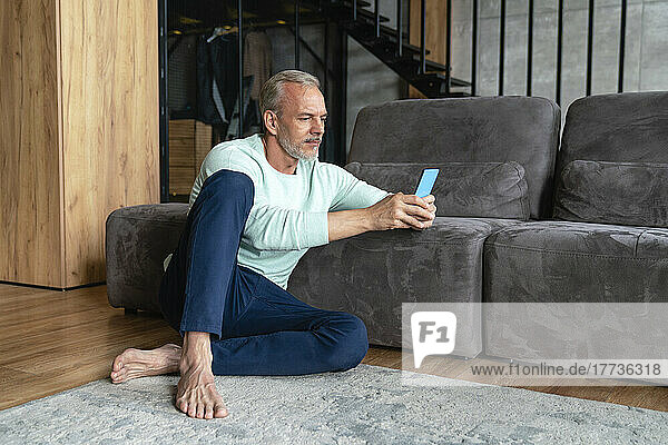 Mature man using mobile phone sitting by sofa at home
