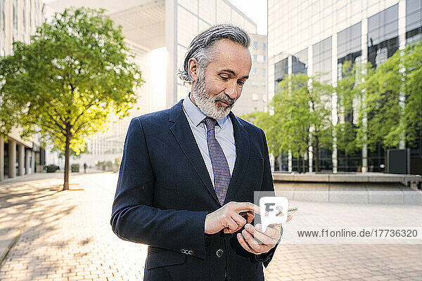 Mature businessman using smart phone standing in office park