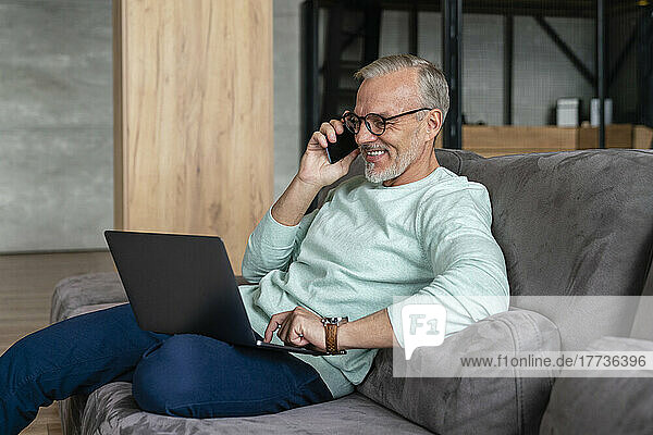 Happy businessman holding laptop talking on mobile phone at home