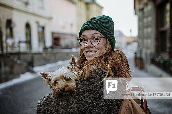 Happy redhead woman wearing knit hat carrying Yorkshire Terrier in city