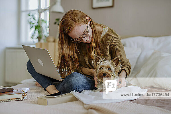 Happy woman wearing eyeglasses stroking Yorkshire Terrier on bed at home
