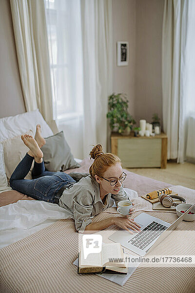 Smiling woman with coffee cup using laptop lying on bed at home