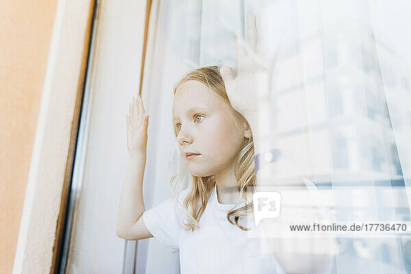 Contemplative girl looking through glass window at home