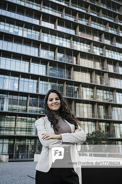 Smiling young businesswoman standing with arms crossed in front of modern office building