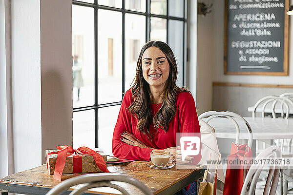 Happy young woman sitting with coffee cup and Christmas present at table in cafe