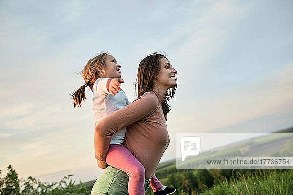 Happy mother piggybacking cute daughter in field at sunset