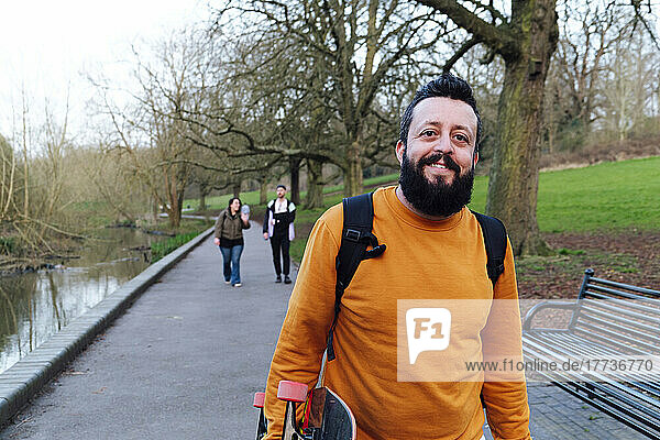 Smiling man carrying skateboard with friends walking in background at park