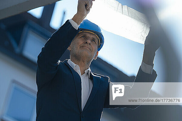 Senior businessman wearing hardhat analyzing blueprint at construction site