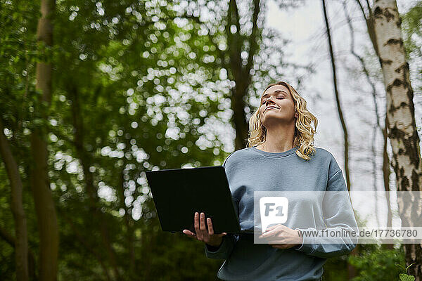 Smiling freelancer with eyes closed holding laptop in forest