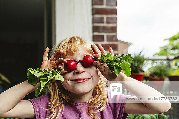 Smiling girl covering eyes with fresh radish on balcony