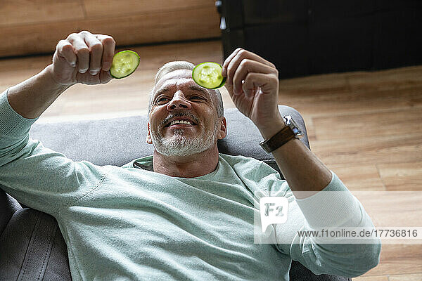 Smiling man looking at slices of cucumber lying on sofa