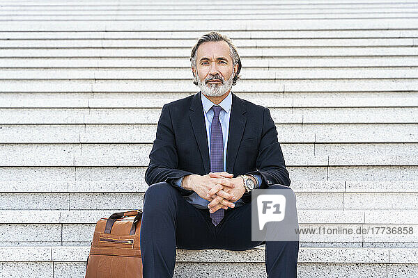 Mature businessman with hands clasped sitting by bag on staircase