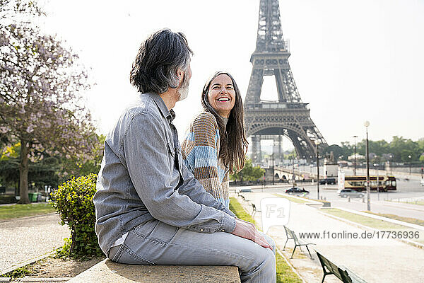 Cheerful mature woman sitting with boyfriend on wall in front of Eiffel tower  Paris  France
