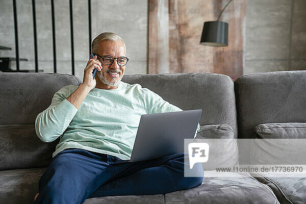 Lächelnder Geschäftsmann hält Laptop in der Hand und telefoniert zu Hause