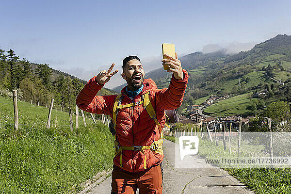Glücklicher Mann gestikuliert Friedenszeichen und macht an einem sonnigen Tag ein Selfie mit dem Smartphone