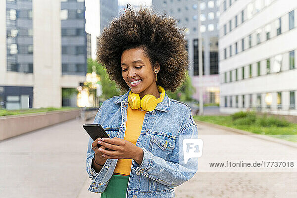 Smiling Afro woman text messaging through smart phone standing on footpath