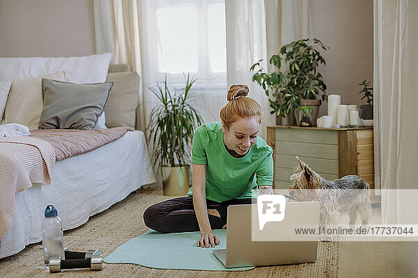 Lächelnde Frau schaut sich Online-Tutorial über Laptop an  während sie zu Hause neben ihrem Hund auf der Trainingsmatte sitzt