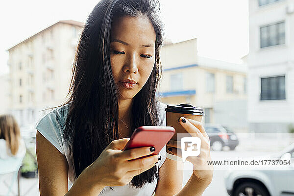 Schöne Frau hält eine wiederverwendbare Kaffeetasse in der Hand und sendet SMS über ihr Smartphone