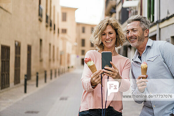 Happy couple with ice cream taking selfie on smart phone in city