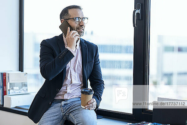 Businessman holding disposable coffee cup talking on smart phone sitting by window in office