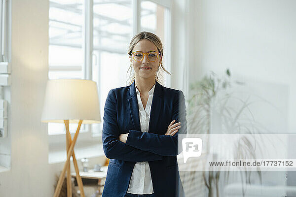 Smiling businesswoman standing with arms crossed in office seen through glass