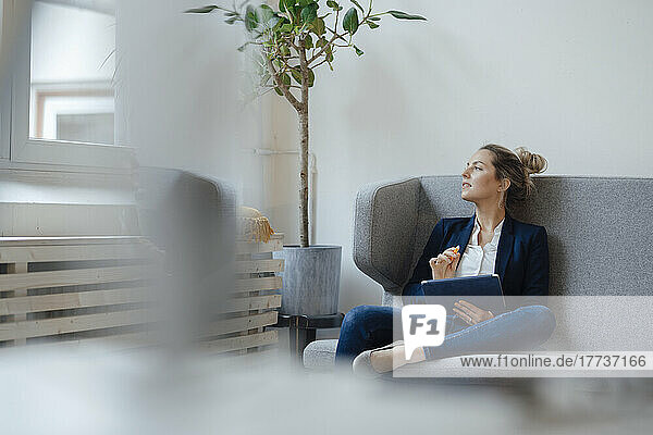 Thoughtful businesswoman holding tablet PC sitting on sofa in office