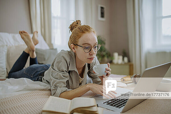 Woman with coffee cup using laptop lying on bed at home