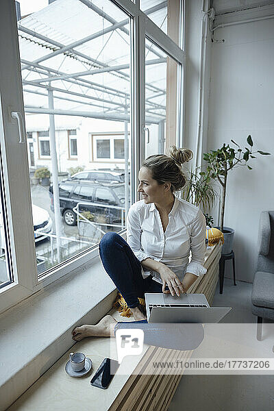 Happy businesswoman with laptop sitting on window sill at home