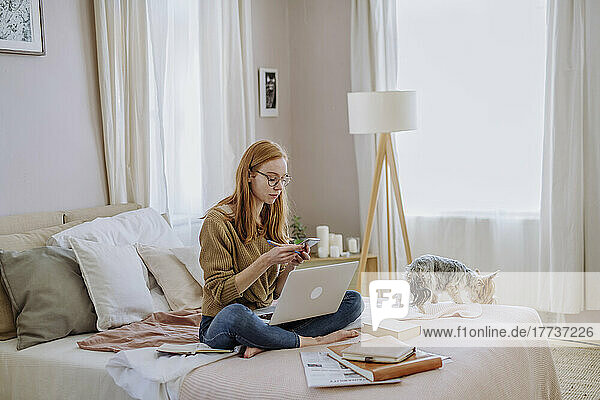 Woman using smart phone sitting with laptop by dog on bed at home