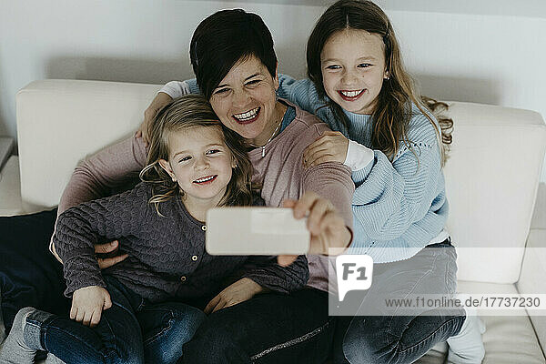 Lächelnde Mutter macht Selfie mit Töchtern  die zu Hause auf dem Sofa sitzen
