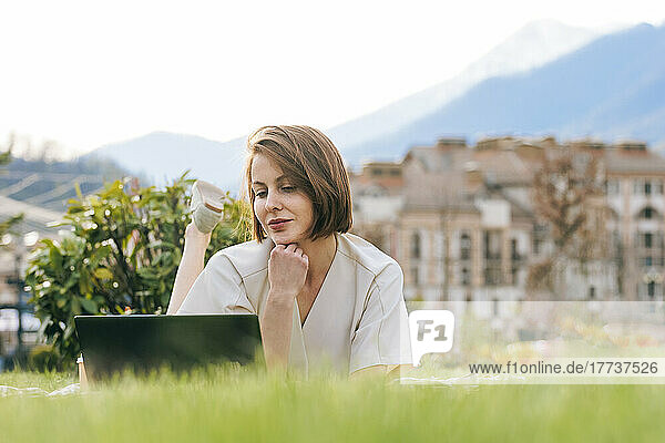 Woman with hand on chin using laptop lying at park