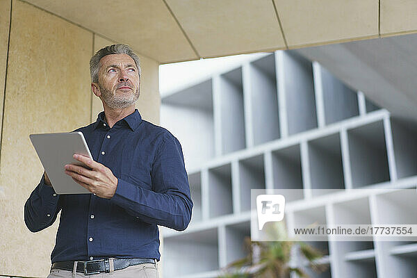 Mature businessman holding tablet PC standing by wall
