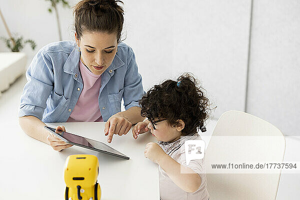 Berufstätige Mutter sitzt am Tisch  Tochter spielt mit KI-Spielzeugroboter