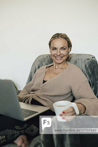 Smiling woman holding coffee cup sitting with laptop on armchair