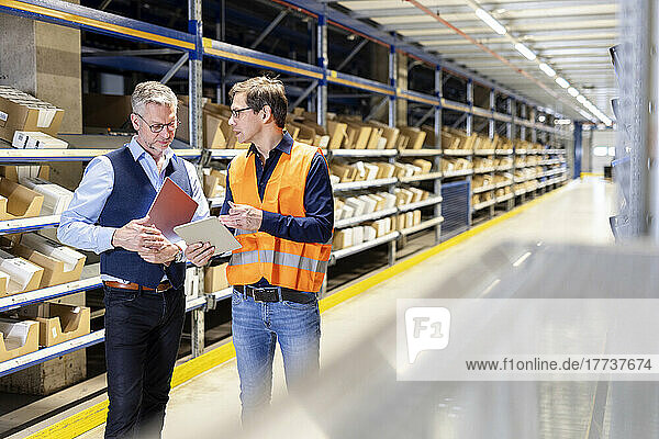 Worker discussing over tablet PC with manager standing in aisle at warehouse