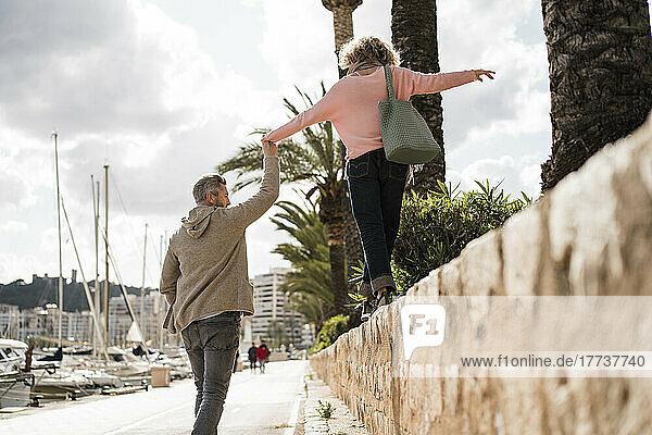 Mature man holding hand of woman walking on wall by harbor