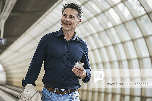 Smiling mature man with mobile phone walking at railroad station