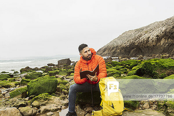 Man with diary and backpack sitting on rock at beach