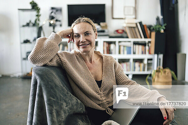 Lächelnde Frau sitzt mit Laptop im Sessel zu Hause
