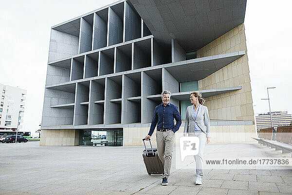 Smiling businessman with luggage walking by businesswoman in front of office building building