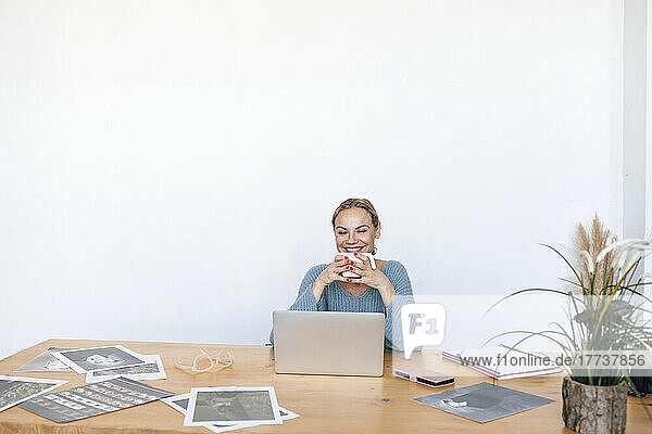 Happy businesswoman holding coffee cup sitting with laptop at home