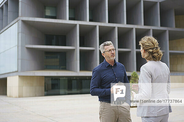 Businesswoman discussing with businessman holding mobile phone standing in front of office building