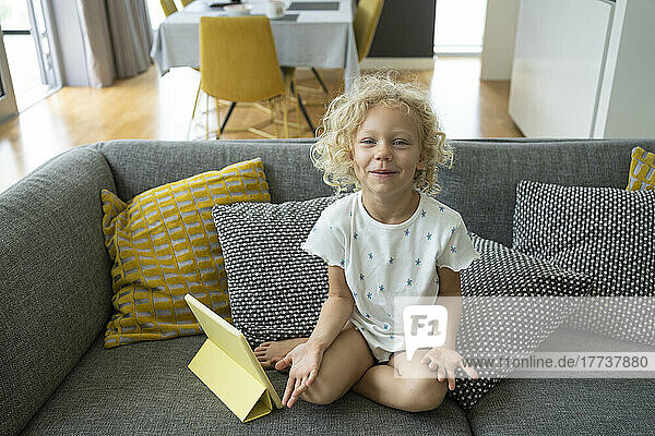 Lächelndes Mädchen mit Tablet-PC sitzt zu Hause inmitten von Kissen auf dem Sofa
