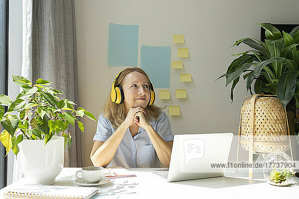 Thoughtful smiling freelancer listening music through wireless headphones sitting with laptop at desk in home office