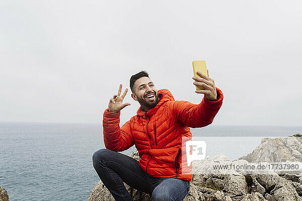 Glücklicher Mann gestikuliert Friedenszeichen und macht ein Selfie mit dem Smartphone  das auf einem Felsen sitzt