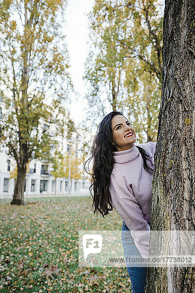 Happy young beautiful woman enjoying autumn standing behind tree trunk at park