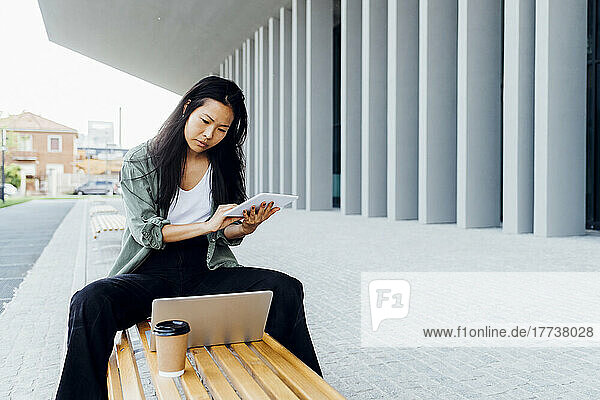Young freelancer wit tablet PC looking at laptop sitting on bench
