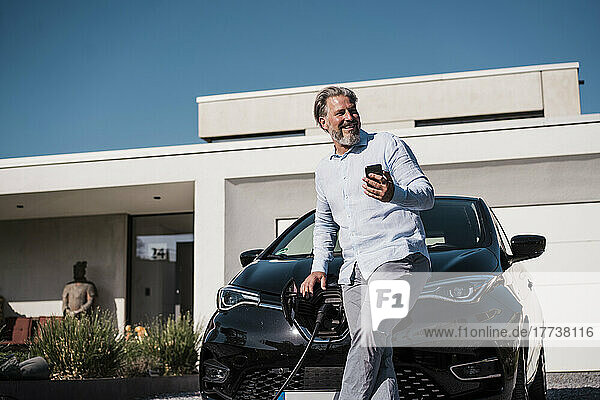 Smiling man holding mobile phone in front of electric car on sunny day