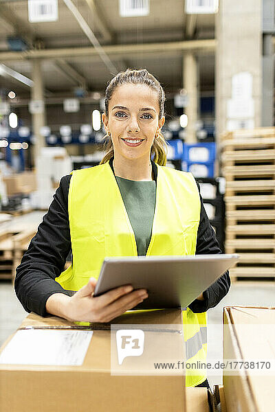 Smiling young worker with tablet PC standing by cardboard box in warehouse