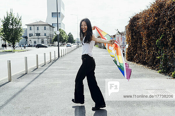 Cheerful young woman with rainbow flag walking on road